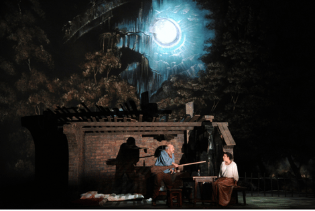 Bühnenbild mit zwei Personen vor einer Ruine unter einem großen, hellen Vollmond und düsteren Bäumen im Hintergrund