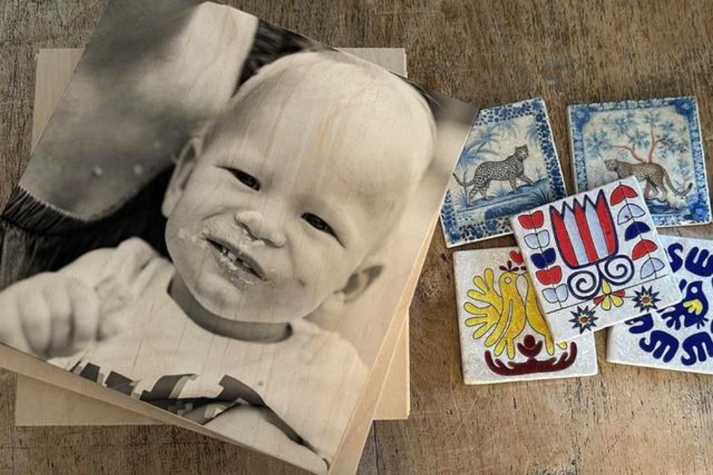Schwarz-weiße Fototransferabbildung eines lächelnden Babys auf Holzplatte neben mehreren bemalten Fliesen mit Tier- und Blumenmotiven auf Holztisch