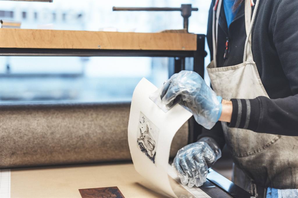 Person mit Schürze und Handschuhen hebt eine Radierung vom Drucktisch ab