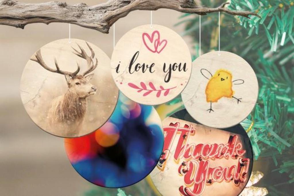 Fünf runde Weihnachtsanhänger mit verschiedenen Motiven hängen an einem Ast, darunter ein Hirsch, Schriftzüge 'i love you' und 'Thank You', ein gelbes Küken und ein bunter abstrakter Kreis