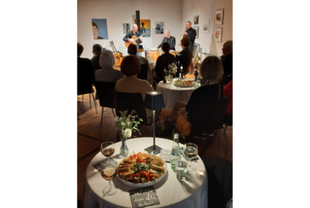 Kleiner Konzertsaal mit Publikum an Tischen, im Hintergrund spielen drei Musiker mit Gitarre und Kontrabass