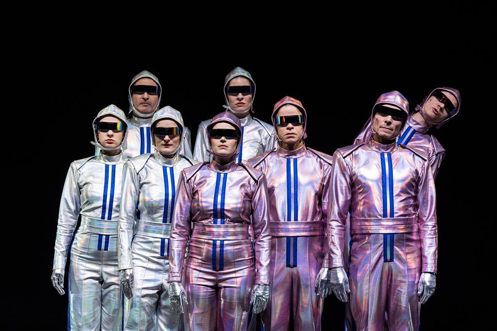 Eine Gruppe von acht Schauspieler*innen steht in silbergrauen und silberrosafarbenen Overalls vor einem schwarzen Hintergrund und blickt durch schwarze Sonnenbrillen Richtung Betrachter*in. 