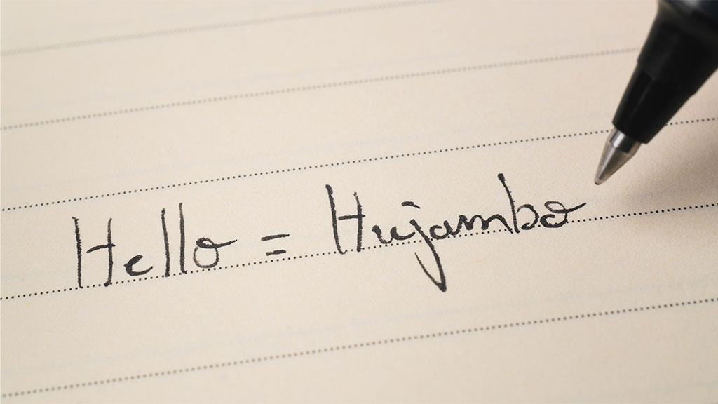 Eine Hand schreibt das Wort ‚Hallo‘ auf Swahili auf ein Blatt Papier