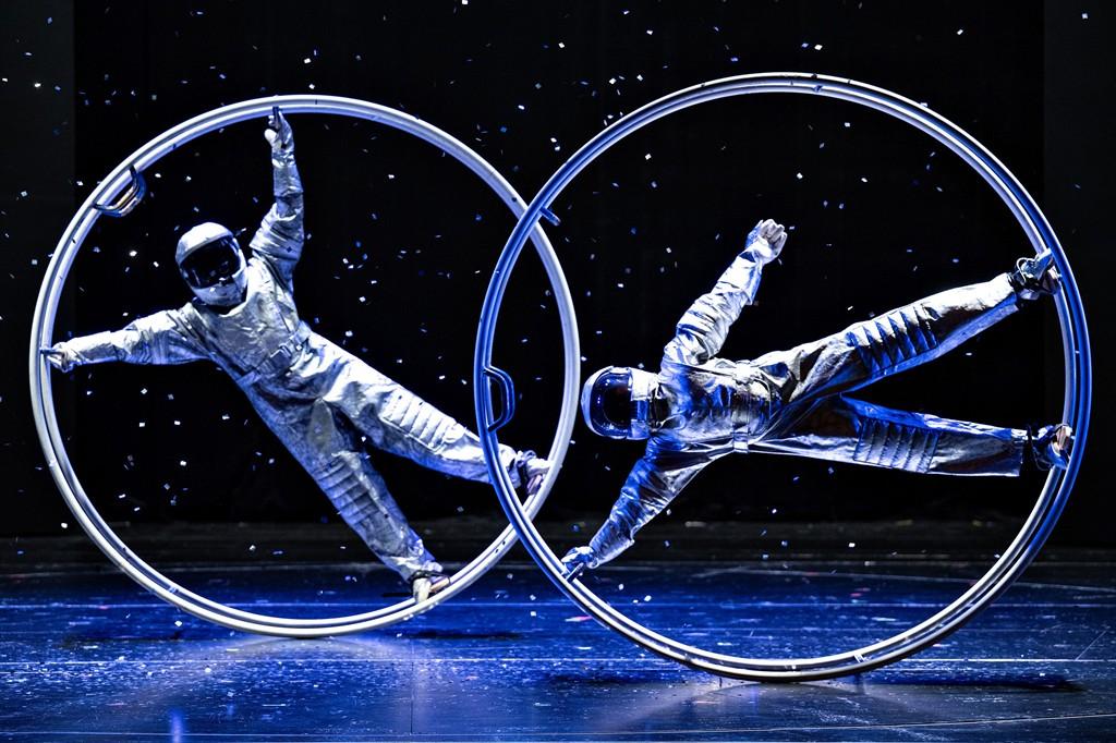 Zwei Artistinnen oder Artisten in silbernen Astronautenanzügen zeigen eine Monorad-Akrobatik auf einer dunklen Bühne im Weltraum-Look.